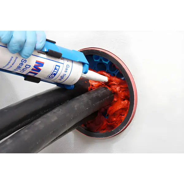Blue caulking gun applying red sealant around black pipes for Filoform Filoseal+HD Fire