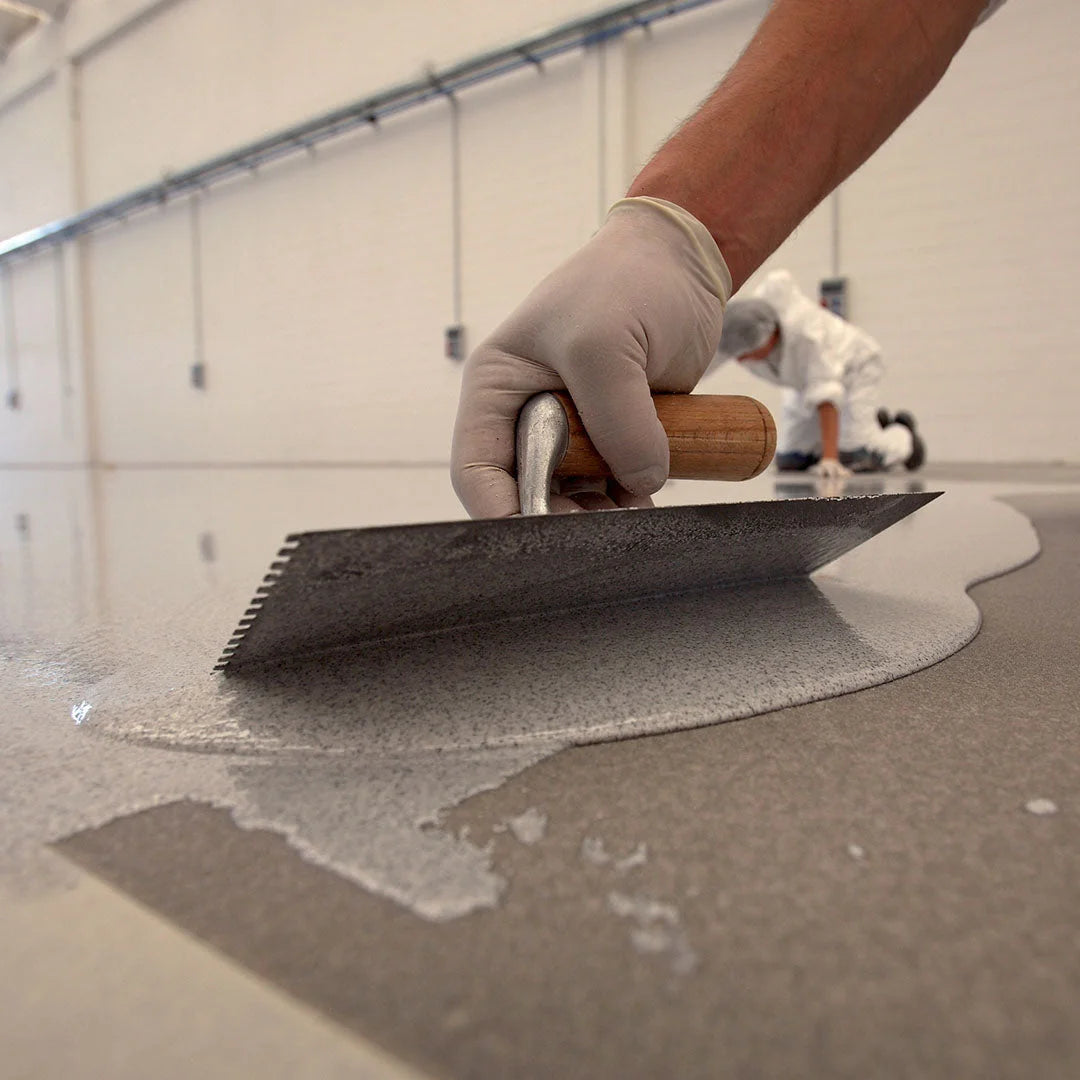 Close-up of MAPEFLOOR I 320 SL CONCEPT being applied with a notched trowel on a concrete floor in an industrial setting