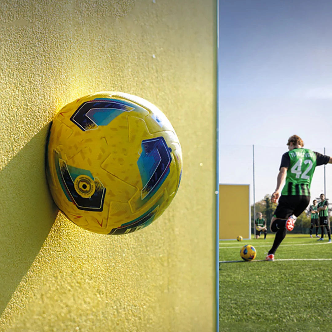 A football hitting a yellow wall with a player preparing for a shot in the background, illustrating the strength and durability of impact-resistant products like Mapetherm Flex RP.

